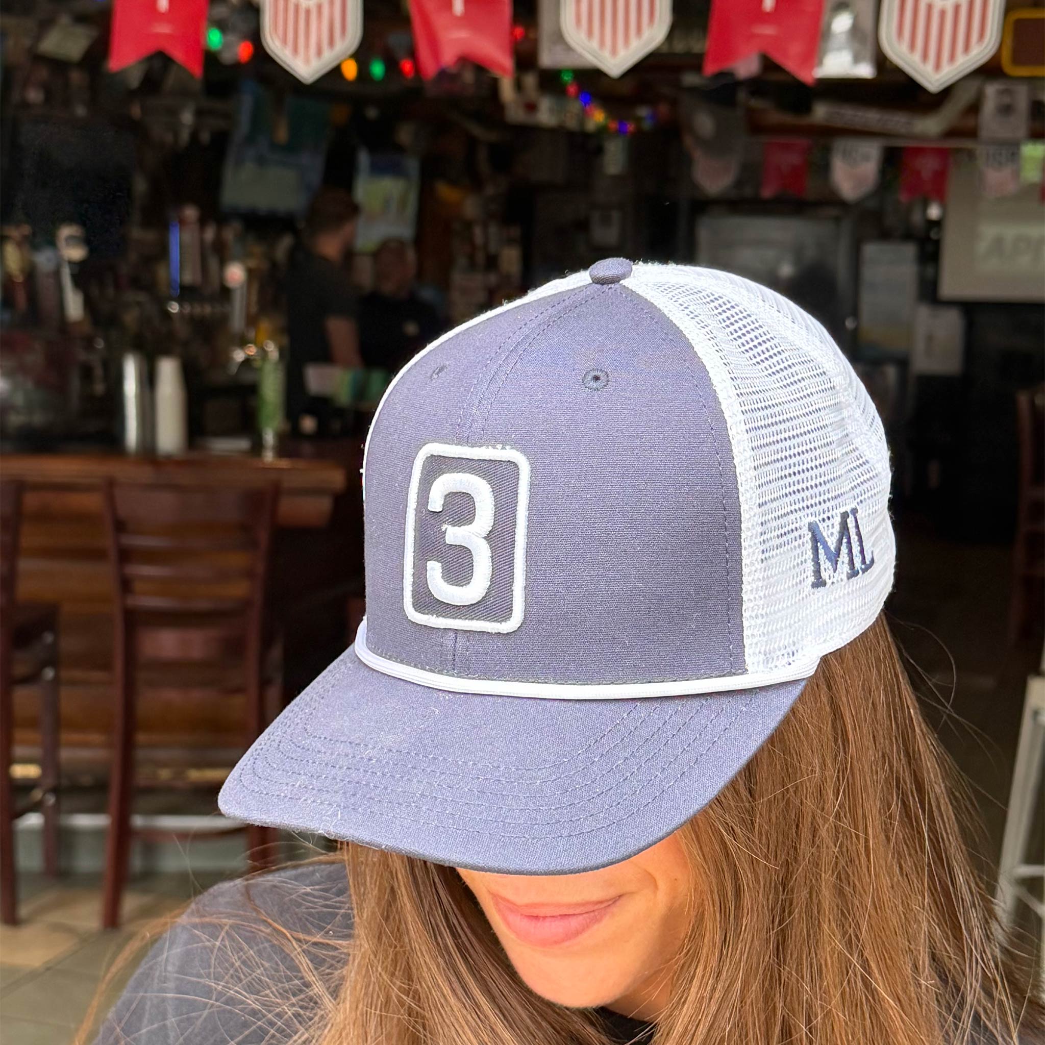 A person wearing a lavender and white trucker hat with a large white '3' on the front panel and 'ML' on the side mesh, photographed in a bar setting.