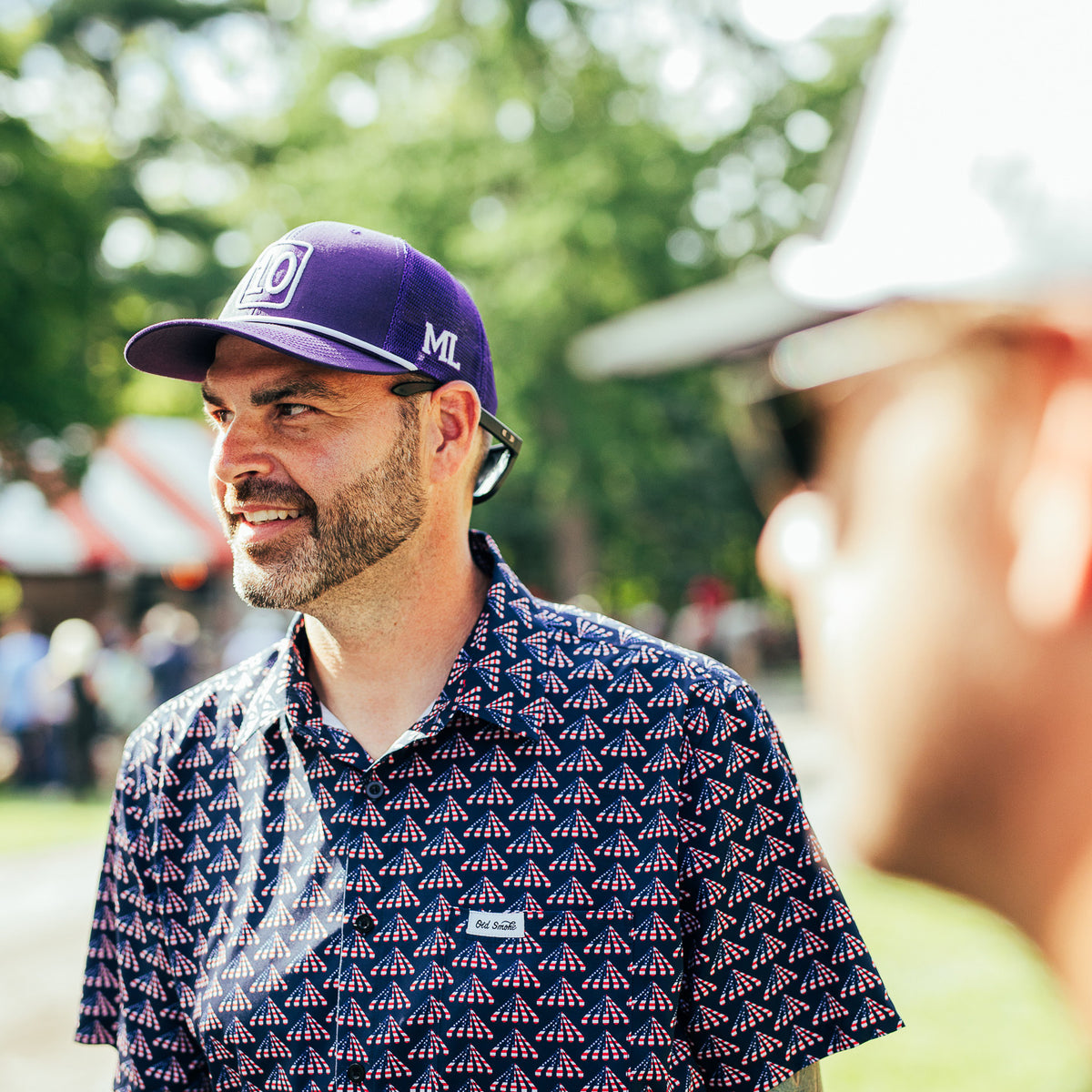 A man outdoors wearing a purple baseball cap with a white 'ML' logo and a navy short-sleeve button-up shirt with a repeating red and white tent-like pattern. Another person is blurred in the foreground, and trees are visible in the background.