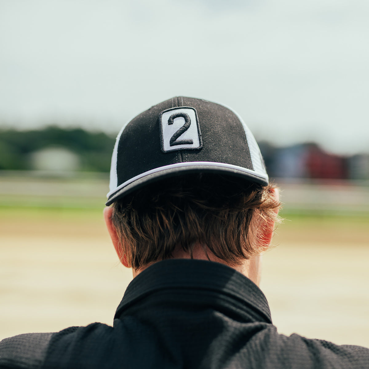 A person viewed from behind wearing a black and white trucker-style baseball cap with a white square patch featuring the number 2, standing outdoors in a blurred field or track setting.