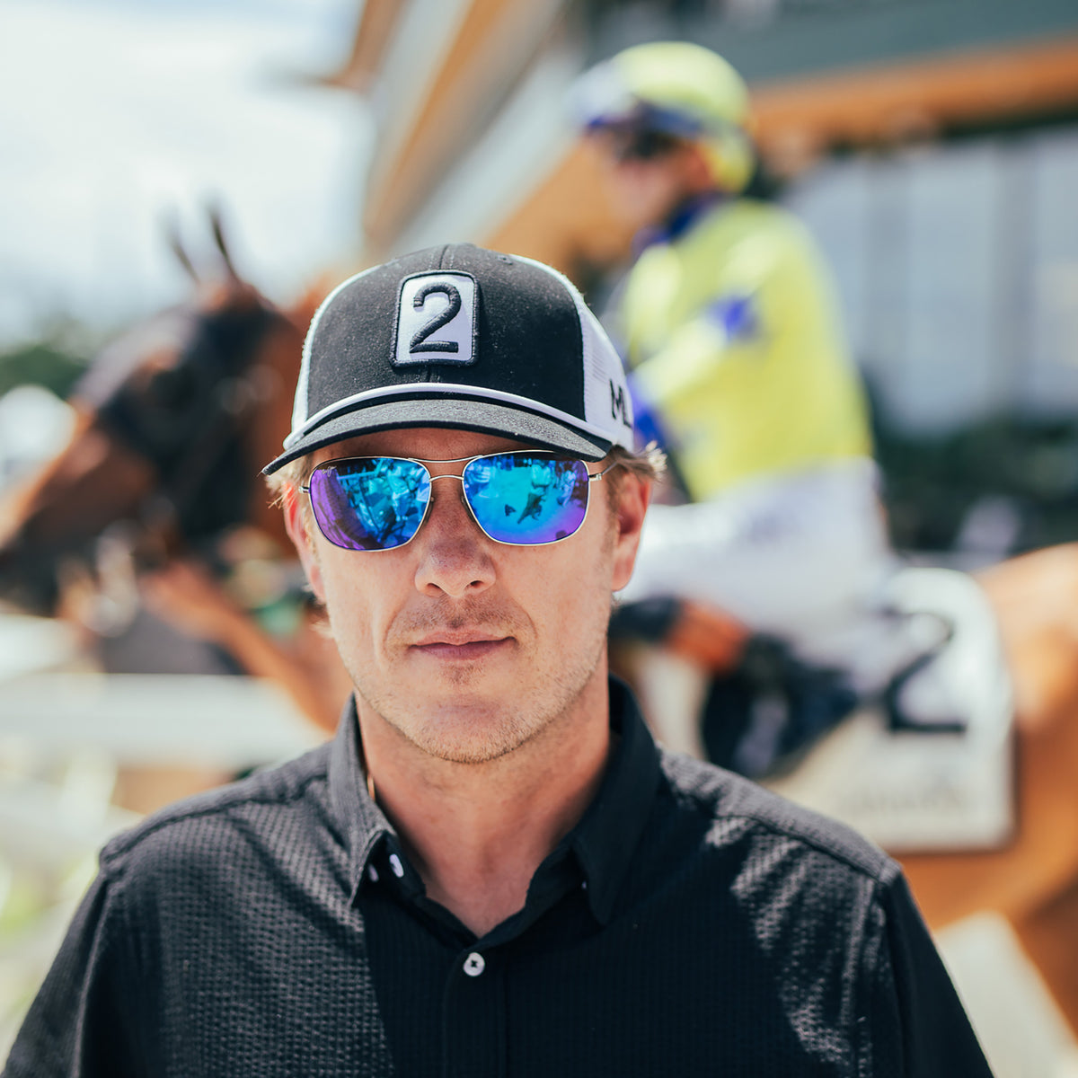 A man in a black shirt wearing a black and white cap with a white number 2 patch and reflective blue-tinted sunglasses, standing in front of a blurred horse racing scene with another jockey and horse in the background.