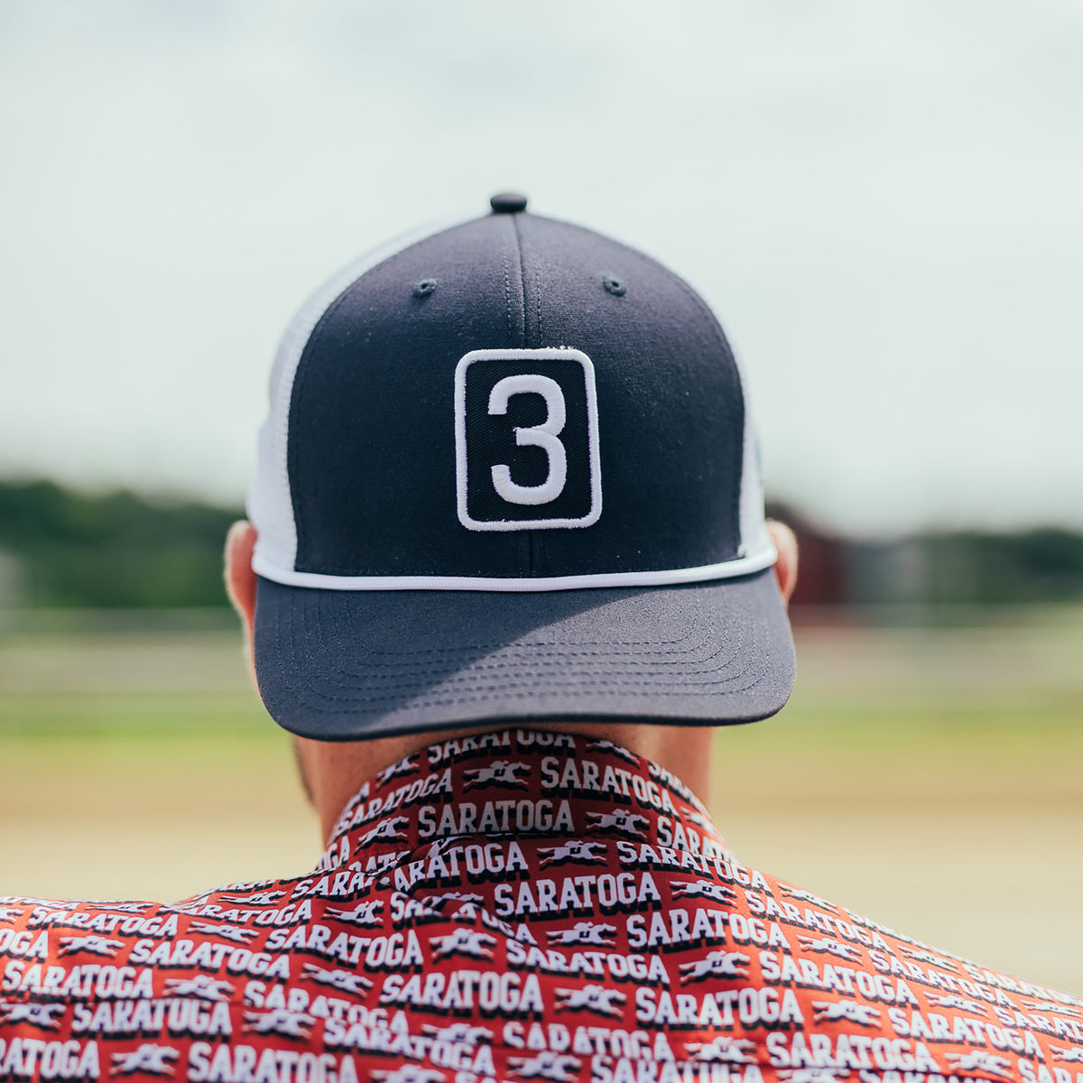 A man viewed from behind wearing a dark navy and white trucker hat with a white outlined number 3 on the front, and a red patterned shirt with the word SARATOGA repeated across it, against an outdoor blurred background.