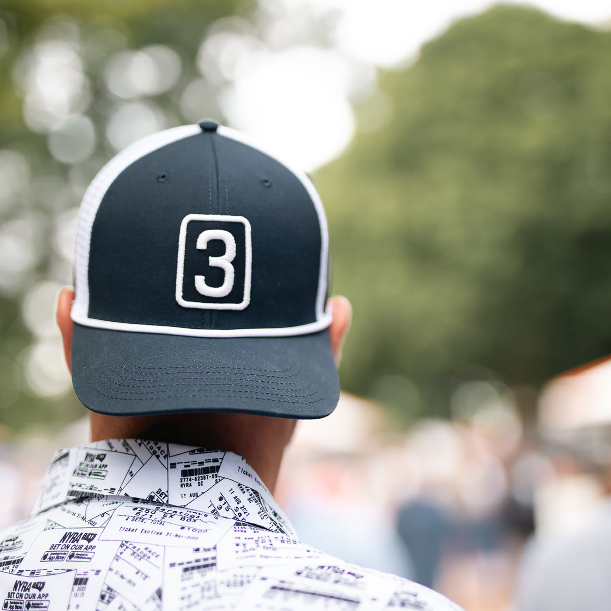 A man viewed from behind wearing a dark navy and white trucker hat with a white outlined number 3 on the front, paired with a white patterned button-down shirt, set against a blurred outdoor background.