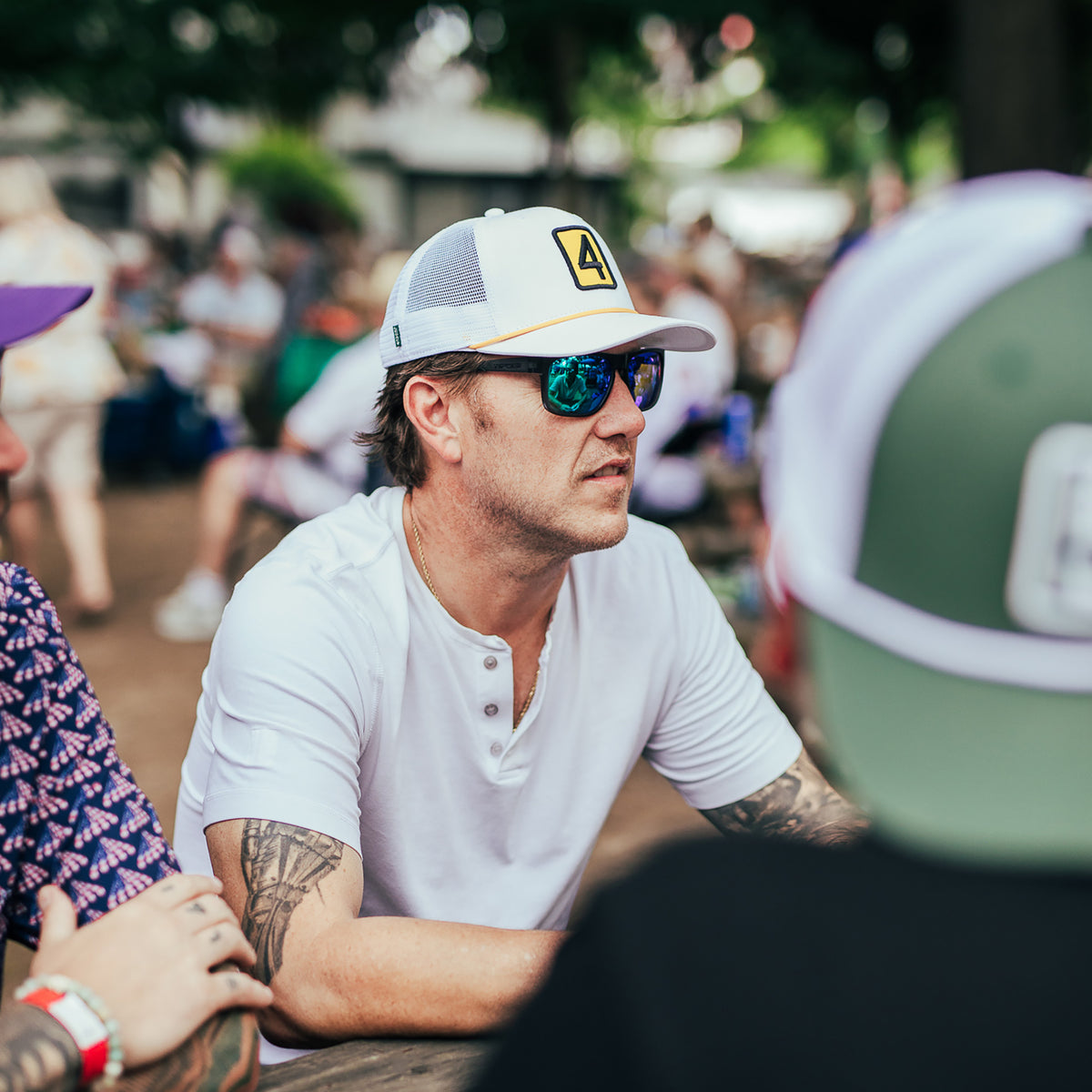 A man wearing a white trucker hat with a yellow graphic logo, blue mirrored sunglasses, and a white henley shirt sits at an outdoor table in a crowded setting.