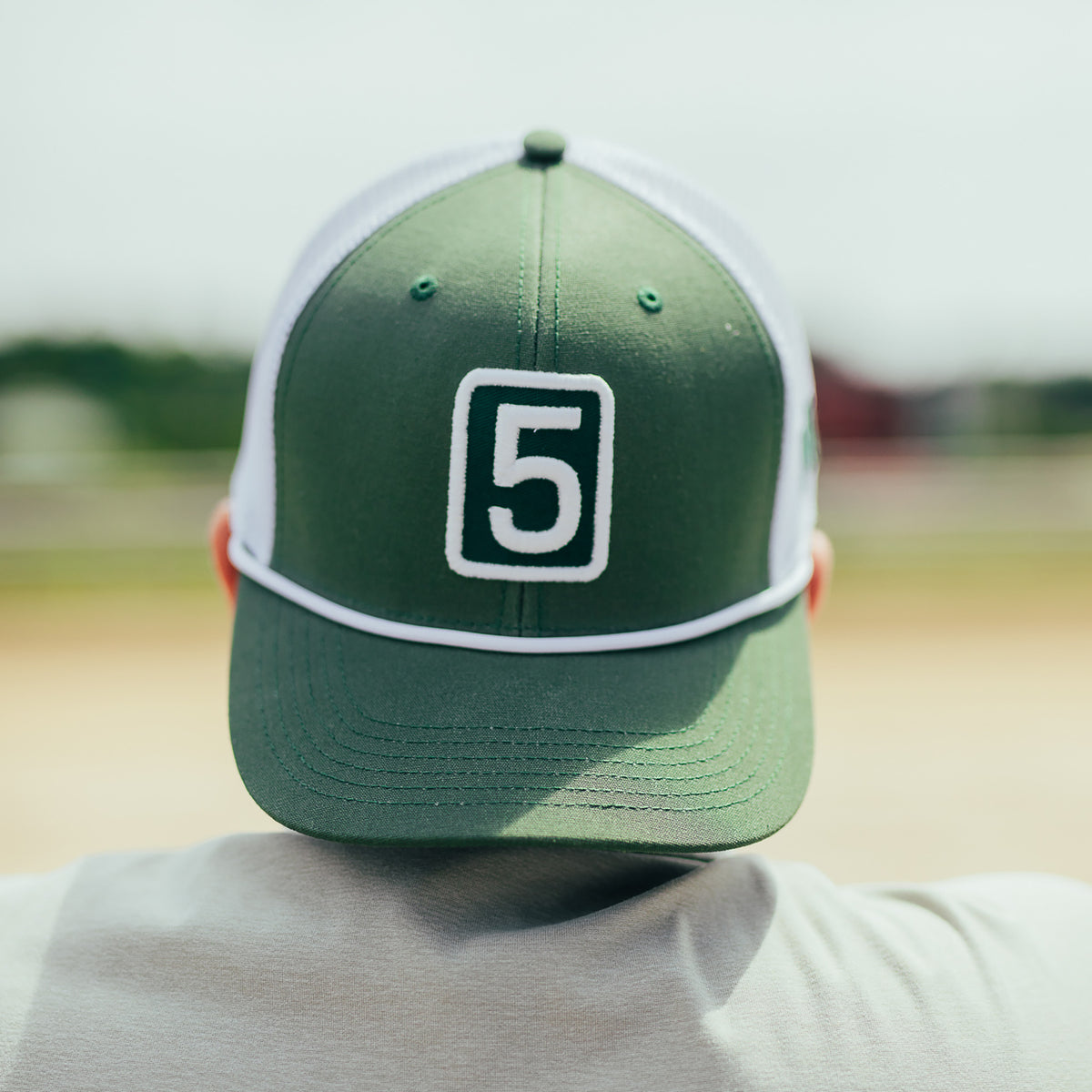 A person viewed from behind wearing a green and white trucker cap with a white outlined number 5 patch on the front, photographed outdoors on a blurred field background.