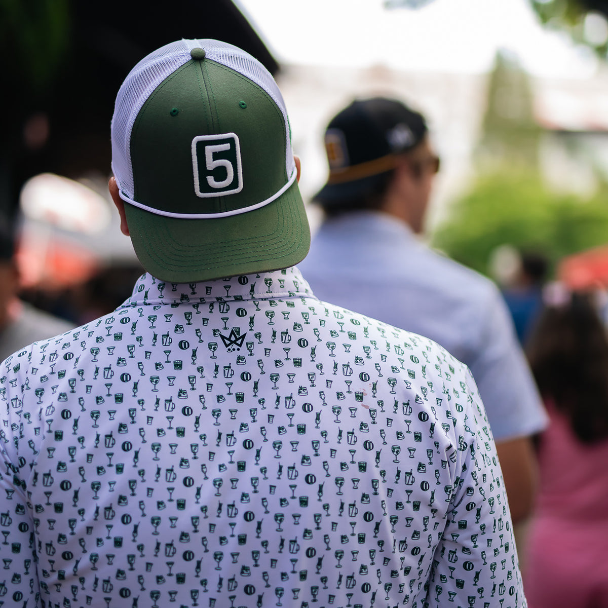A man viewed from behind wearing a green and white trucker hat with a white number 5 patch on the back, and a white short-sleeve button-up shirt covered in a repeating green trophy print. The background is a blurred outdoor crowd scene.