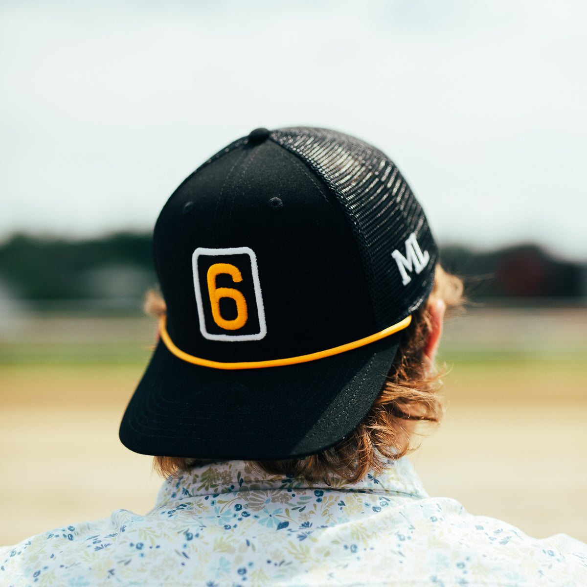 A person viewed from behind wearing a black trucker-style cap with a yellow and white outlined number 6 on the front, a yellow stripe across the brim, and white lettering on the side, set against a blurred outdoor background.