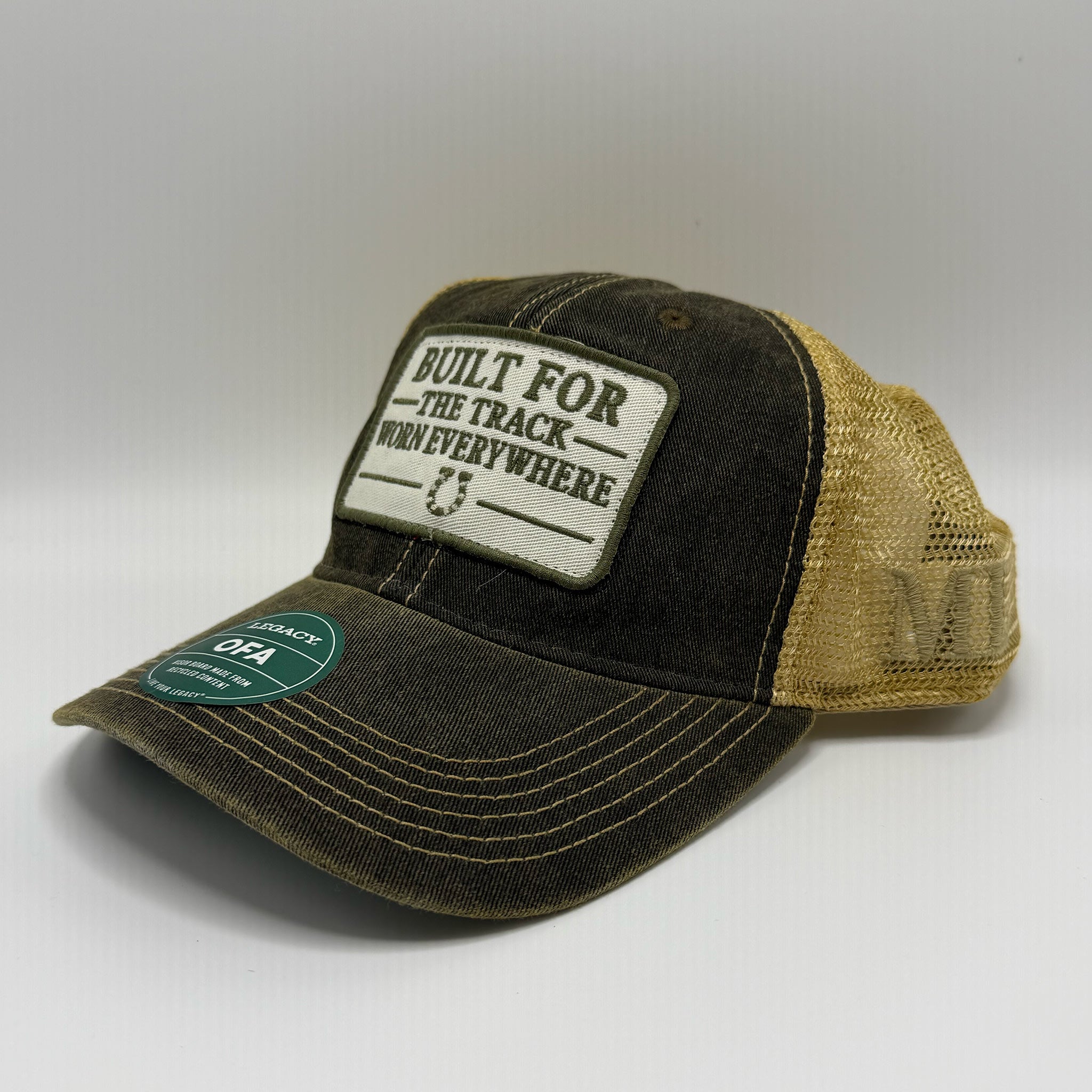 A trucker-style baseball cap with a dark washed front panel and brim, tan mesh back, and a rectangular front patch reading 'BUILT FOR THE TRACK WORK EVERYWHERE.' A green circular size sticker is visible on the brim.